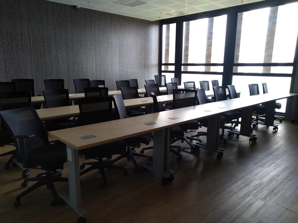 Aula de capacitación universitaria amplia equipada con mesas rectangulares móviles y sillas ergonómicas negras con rodachines. Espacio con grandes ventanales y distribución tipo auditorio.